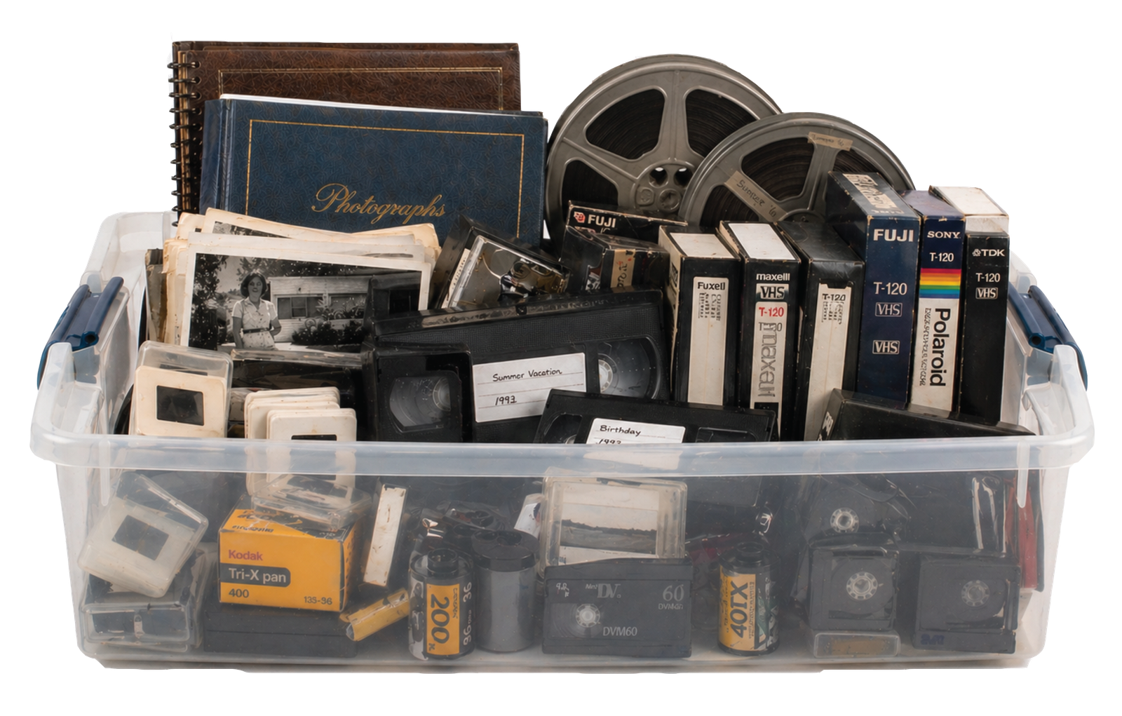 A plastic storage bin overflowing with old family media: photo albums, loose photographs, film reels, VHS tapes, slides, film canisters, and MiniDV cassettes
